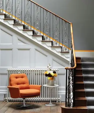 Tromba delle scale verniciata in ardesia con strisce arancioni dipinte, rivestimento del sottoscala verniciato di bianco, scale in legno scuro con passatoia in moquette grigia, pavimento in legno scuro con poltrona lounge arancione brillante, tavolo tulipano bianco con vaso di fiori