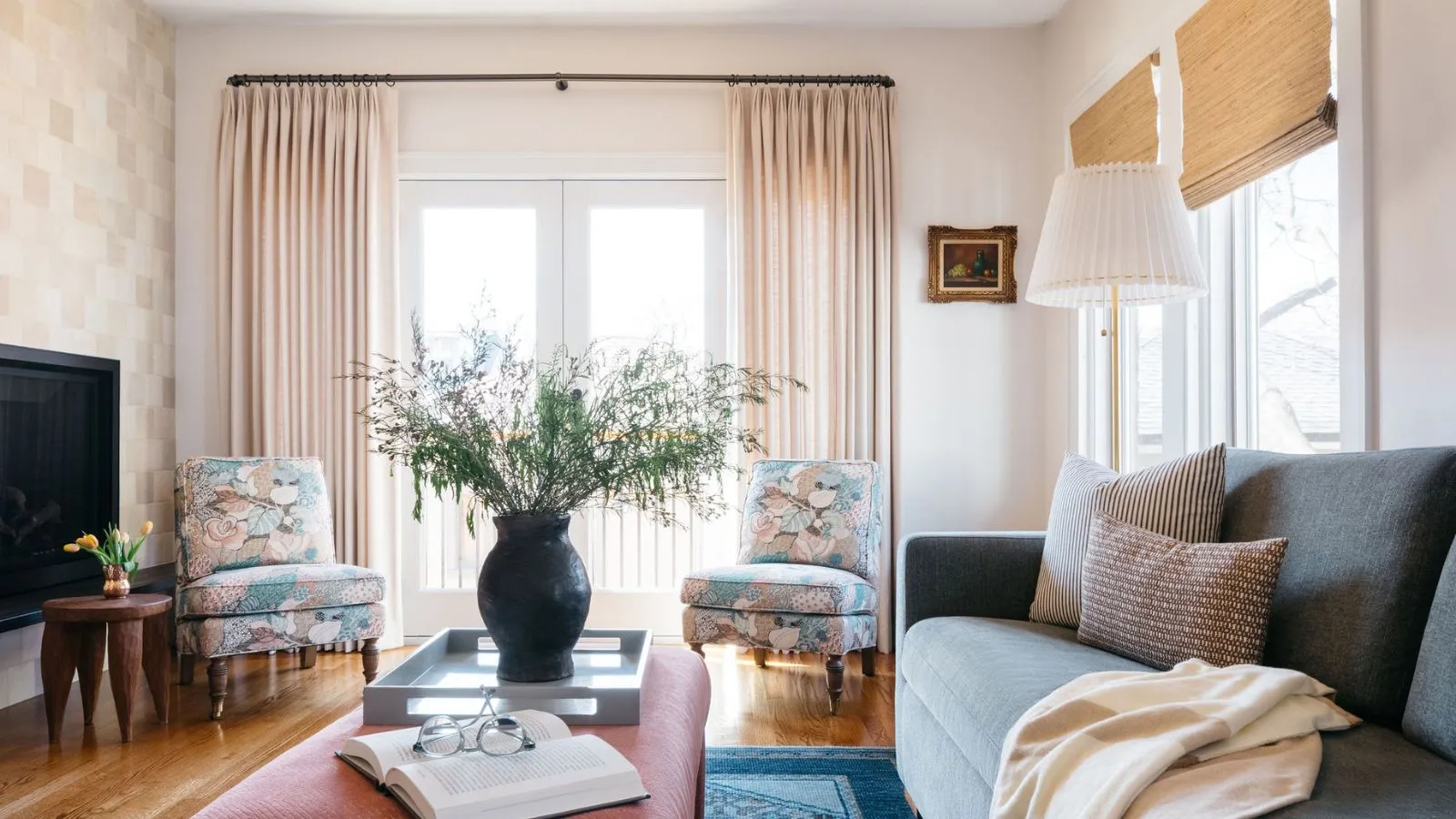 Pokój dzienny z balkonem na ścianie wyłożonej kafelkami, drewnianą podłogą, różowym stolikiem kawowym z tacą i wazonem, fotelami w kwiaty w liście i rattanowymi żaluzjami w oknach' loading='lazy' title=