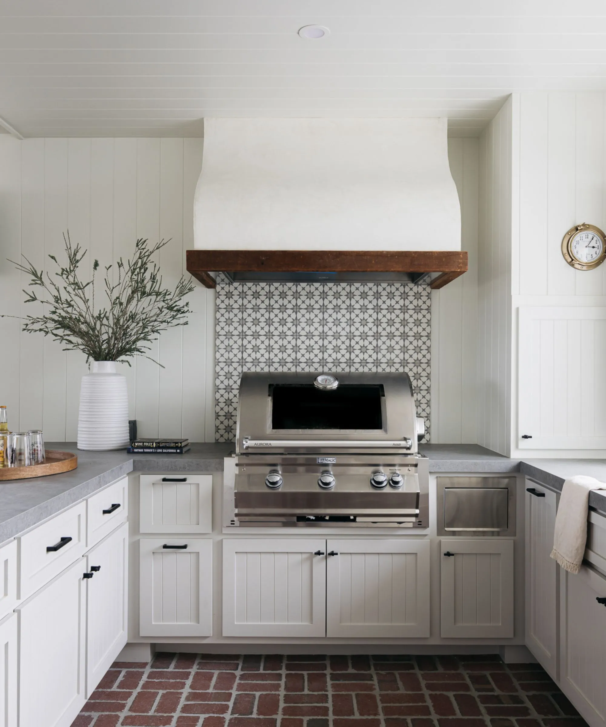 Nowoczesna, rustykalna kuchnia z białawymi szafkami i ścianami, podłogą z cegły i tylną ścianą piekarnika kaflowego.' loading='lazy' title=