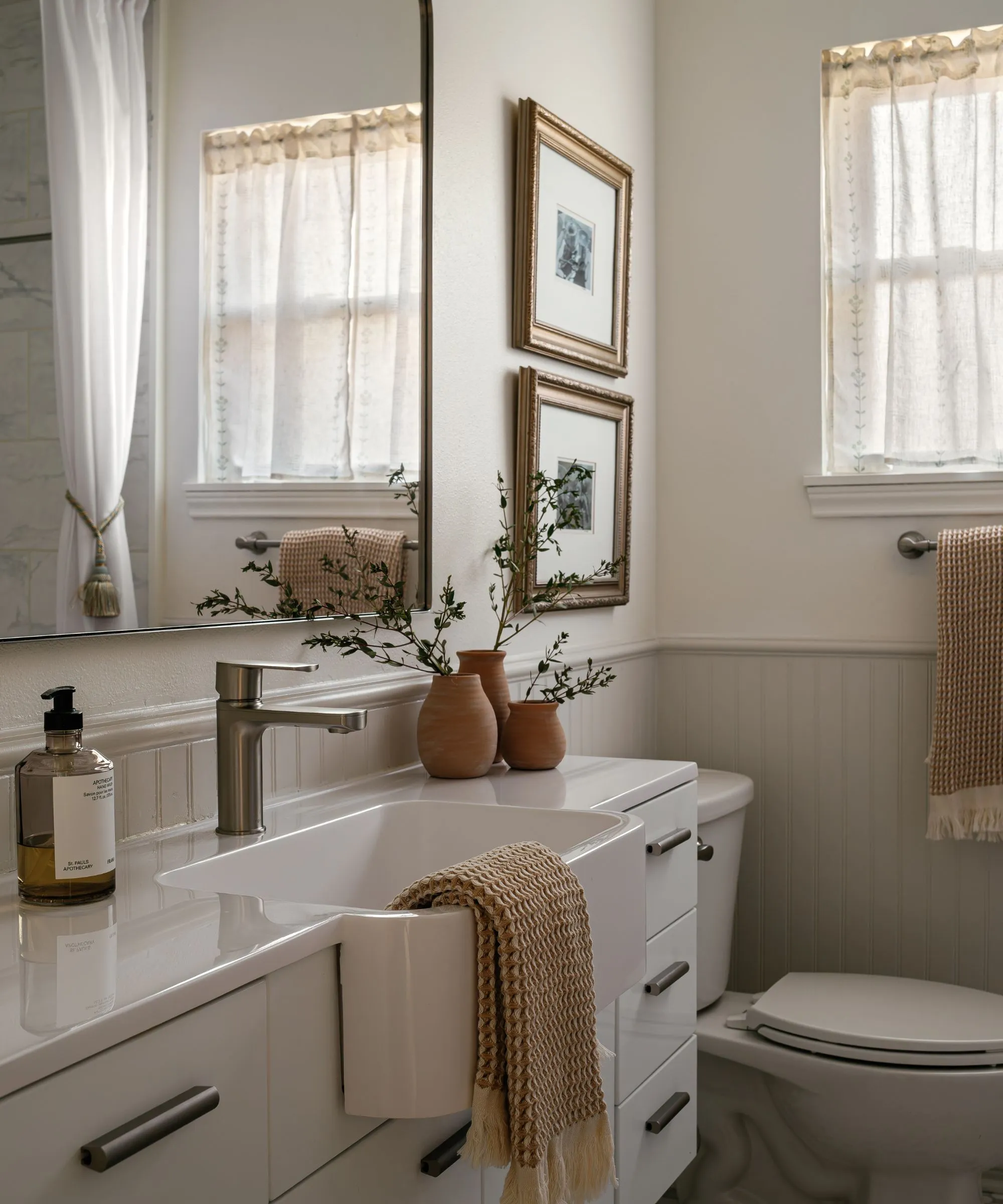 Mała łazienka z jasnoszarymi boazeriami, białymi ścianami powyżej i przytulnym rustykalnym wystrojem, w tym kawiarnianą zasłoną i terakotowymi doniczkami.' loading='lazy' title=