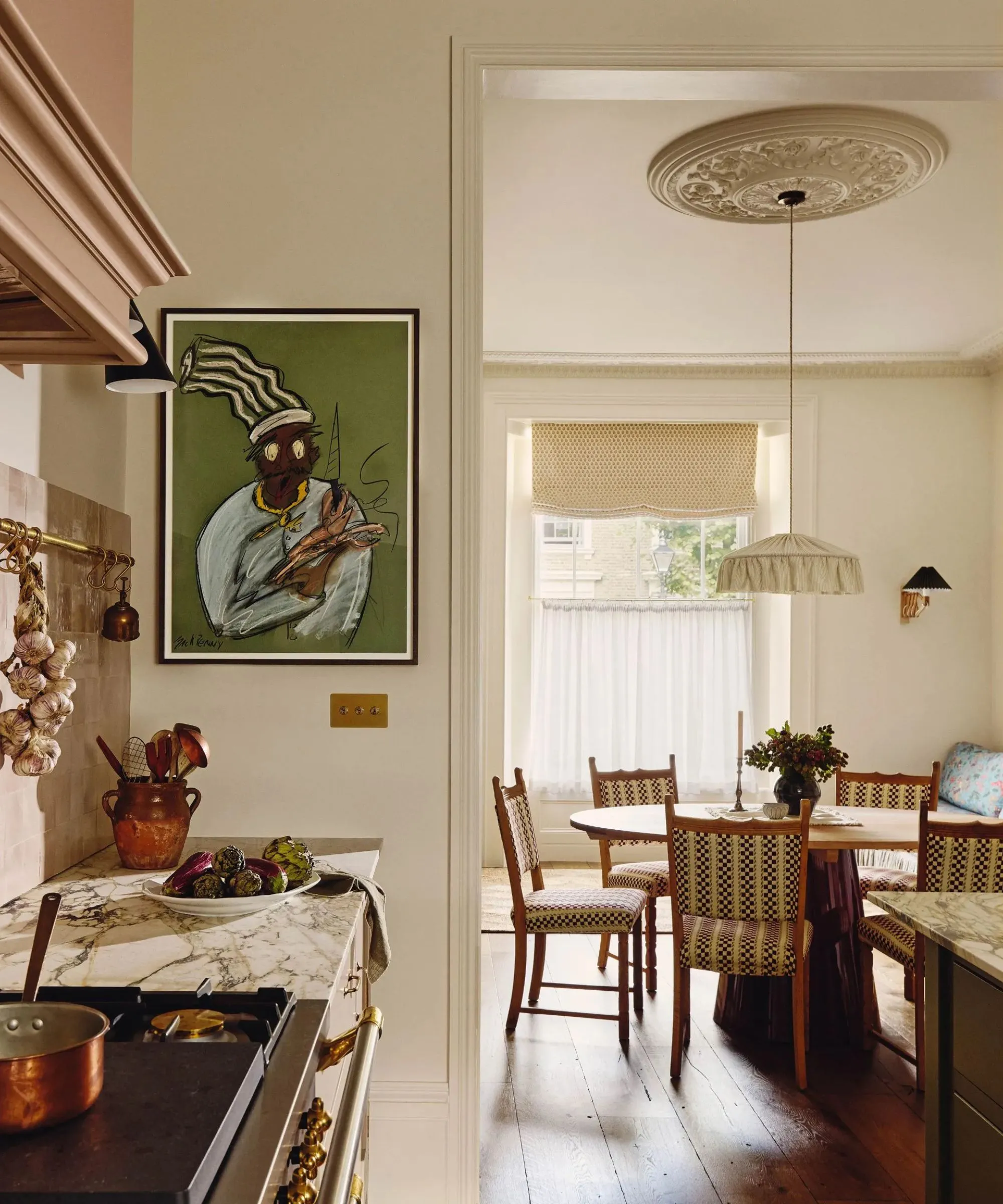 Roh tradiční kuchyně s mramorovou deskou abstraktního umění na stěně a výhledem do přilehlého jídelního prostoru s kulatým dřevěným jídelním stolem a velkým látkovým závěsným světlem nahoře' loading='lazy' title=