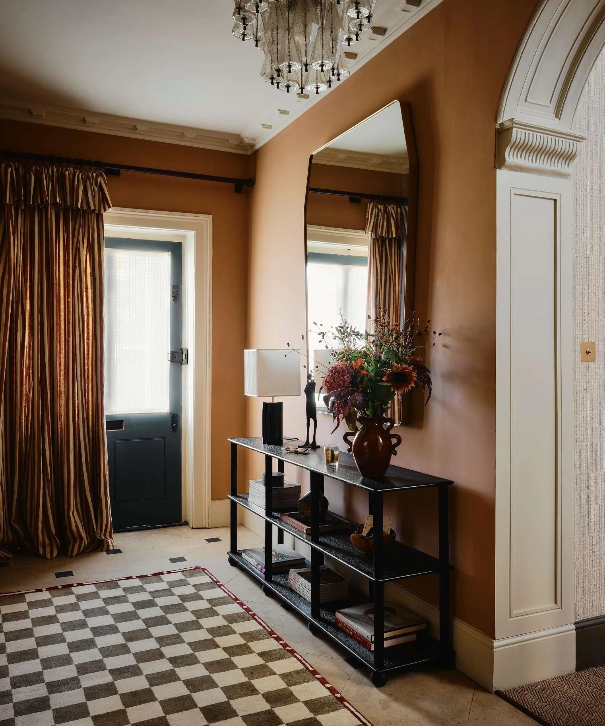 teplá okrová chodba s dlážděnou podlahou šachovnicový koberec velké zrcadlo a konzolový stolek viněta a pruhovaný řasený závěs u předních dveří' loading='lazy' title=