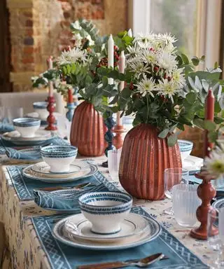 Nahaufnahme eines dekorierten Esstisches mit orange, blau und cremefarben gemusterter Tischdecke, blauen Tischsets aus Stoff, auf dem Tisch liegenden weißen und blauen Tellern und Schüsseln, orangefarbenen Vasen mit Blumen und Grünpflanzen, orangefarbenen Kerzenhaltern mit orangefarbenen, cremefarbenen und rosa Kerzen
