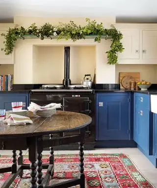 Küche mit cremefarbenen Wänden, leuchtend blauen Schränken, schwarzem Aga mit Regal darüber, verziert mit Blattwerk und Blumen, abgerundetem Esstisch aus dunklem Holz, rotem, blauem und grünem Blumenteppich unter dem Esstisch
