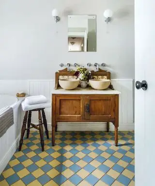 Unidad de madera tradicional con dos lavabos redondos en un baño con baldosas victorianas en azul y amarillo.