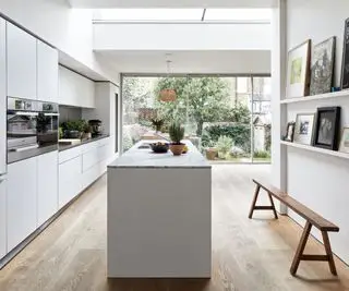 Cocina espaciosa y angular con isla de cocina en blanco, pisos de madera con vista al jardín con puertas francesas y obras de arte en estantes con un banco debajo