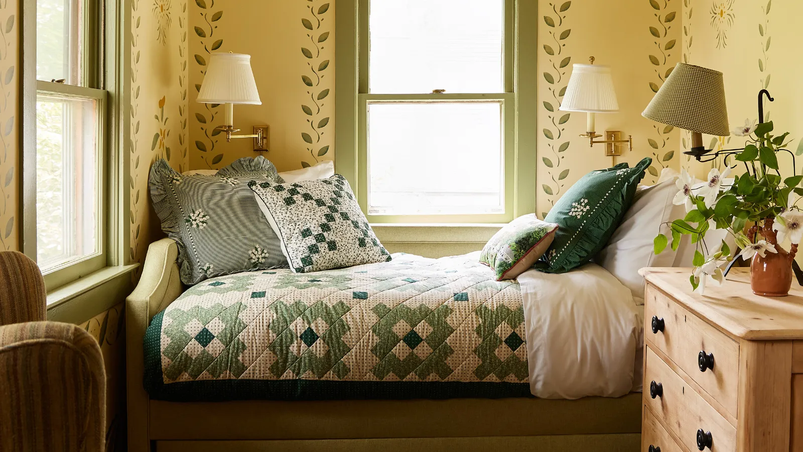 un angolo camera da letto con un letto di patchwork, diversi cuscini un muro di legno e applique e carta da parati giallo con motivi Ivy' loading='lazy' title=