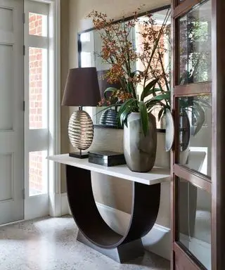 En moderne gangide med stort moderne konsollbord med buet underside, toppet med bordlampe og vase med blomster