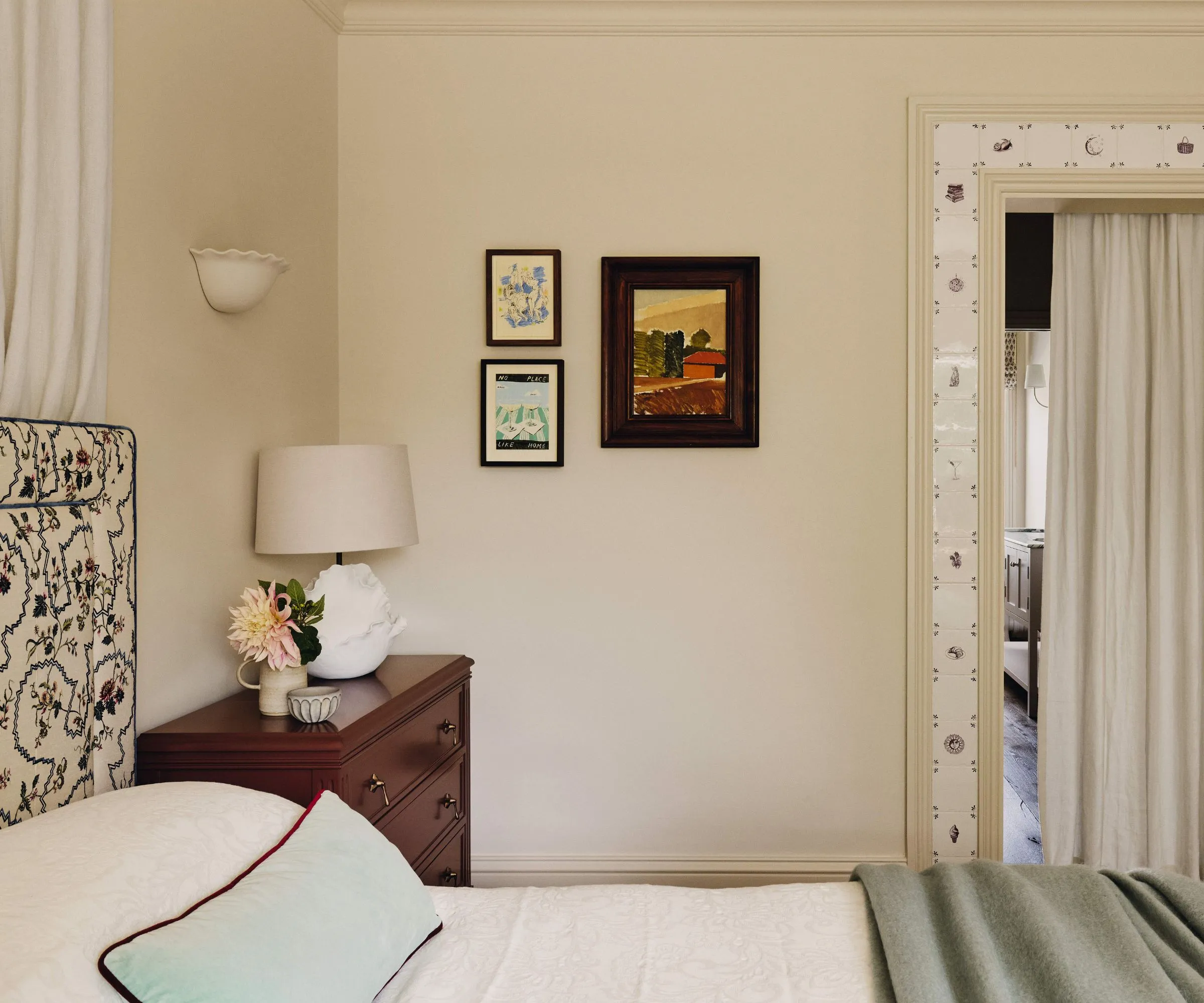 Et kremsoverom med en polstret sengegavl med blomstermønster sammen med et nattbord av tre. På veggen er det tre små innrammede kunstverk. Det er en dør gjennom til badet dekket med et kremgardin som er omgitt av Delft-fliser.' loading='lazy' title=