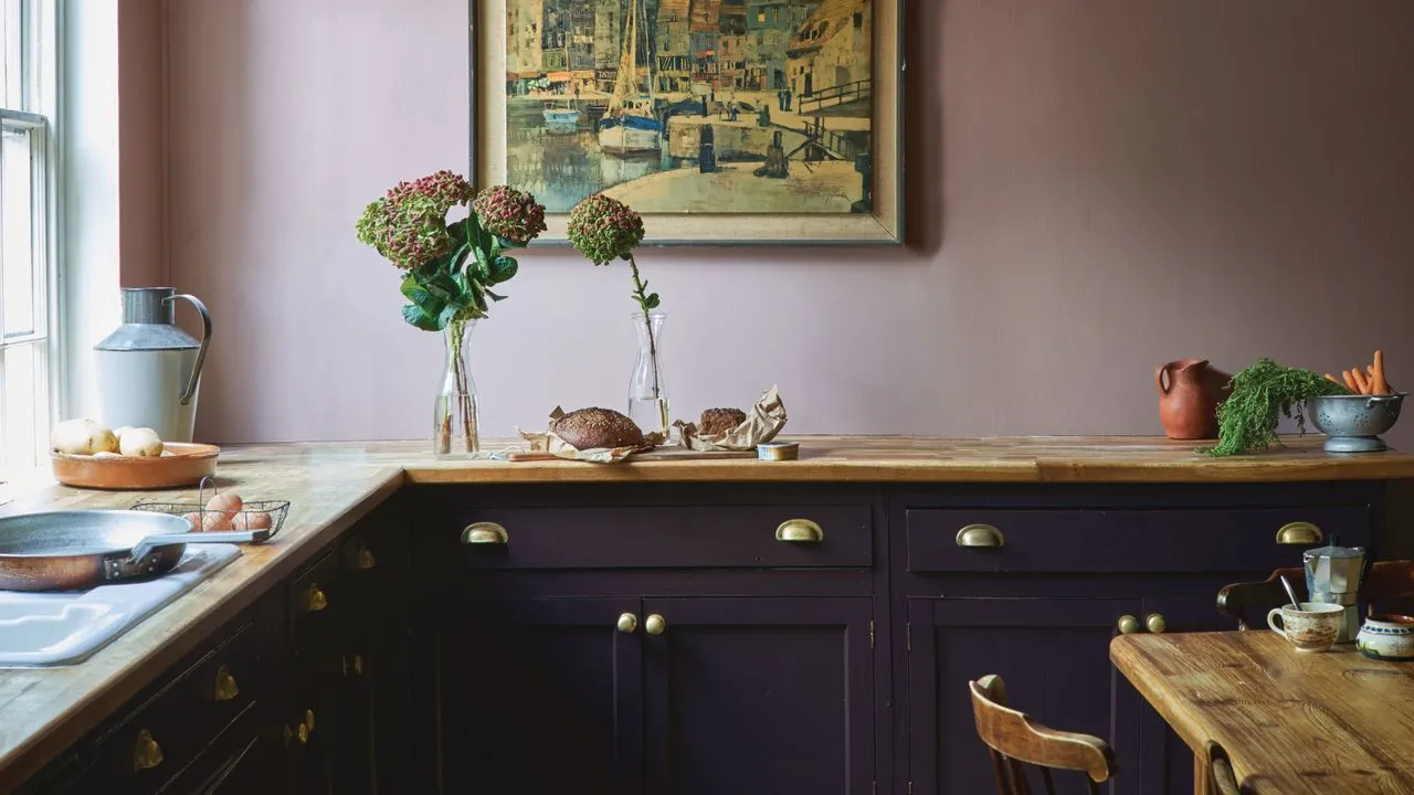Farrow & Ball kjøkken med en rosa vegg og mørk lilla skap med arbeidsflate i tre og rustikk spisebord og stol i tre' loading='lazy' title=