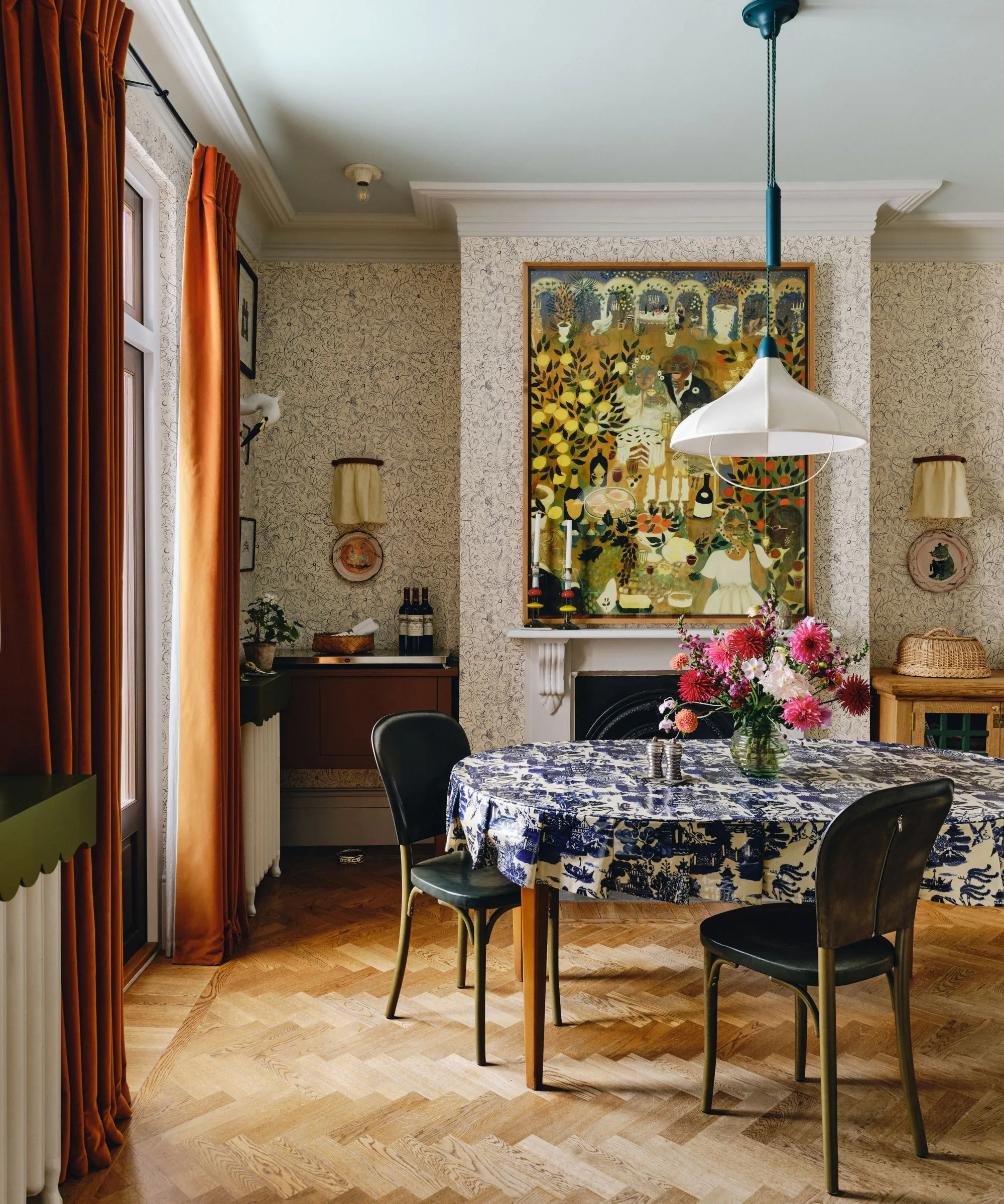 jadalnia w skandynawskim stylu maksymalistycznym ze ścianami wyłożonymi tapetą duży kominek z dziełami sztuki ludowej zawieszonymi nad ciemnopomarańczowymi aksamitnymi zasłonami stół jadalny z niebiesko-białym obrusem z nadrukiem i lampą wiszącą z białej tkaniny' loading='lazy' title=