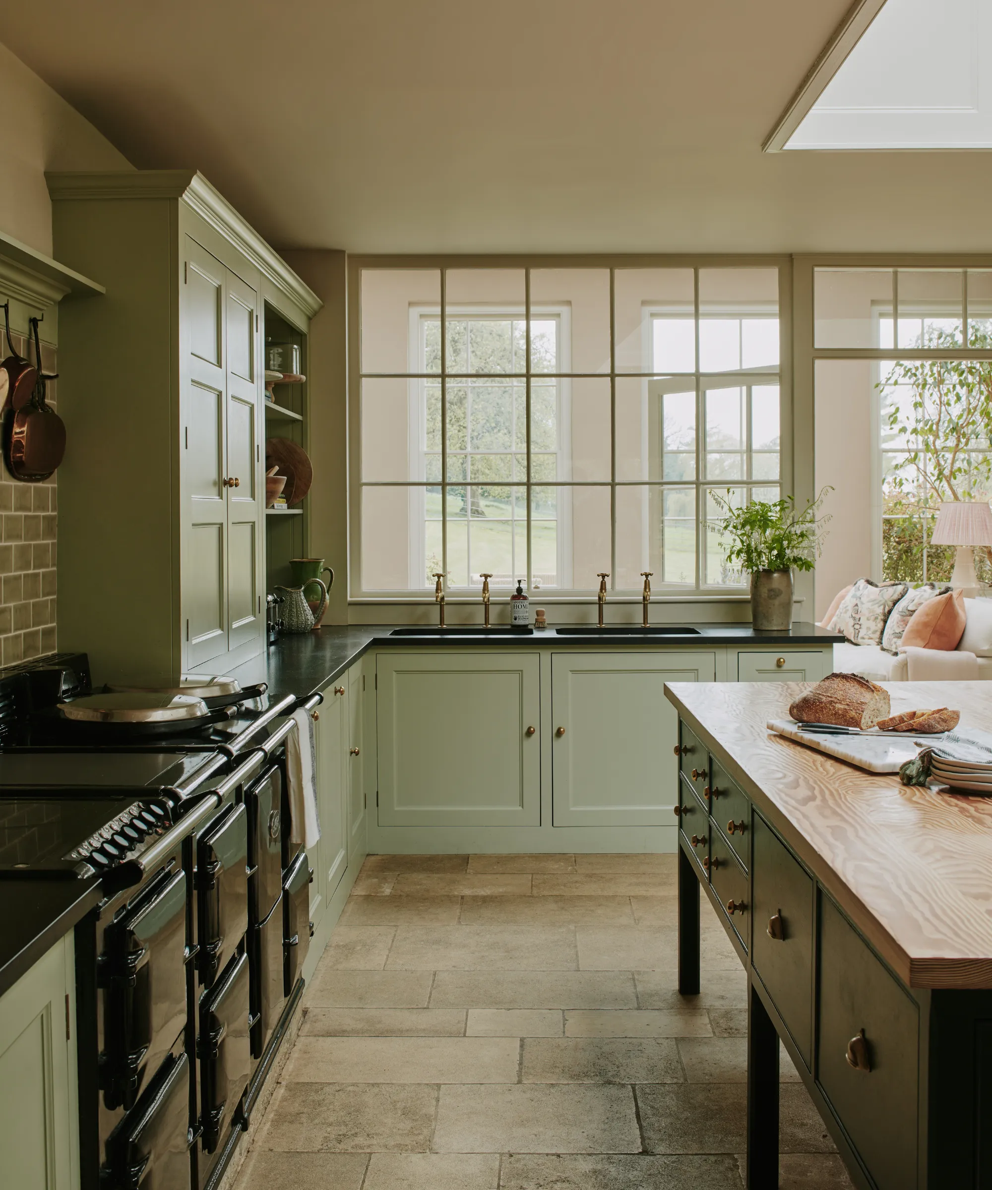 et stort køkken med salviegrønt skab en fritstående ø med en træpladeoverflade en stor ovn og vinduer, der ser ud til en gård' loading='lazy' title=