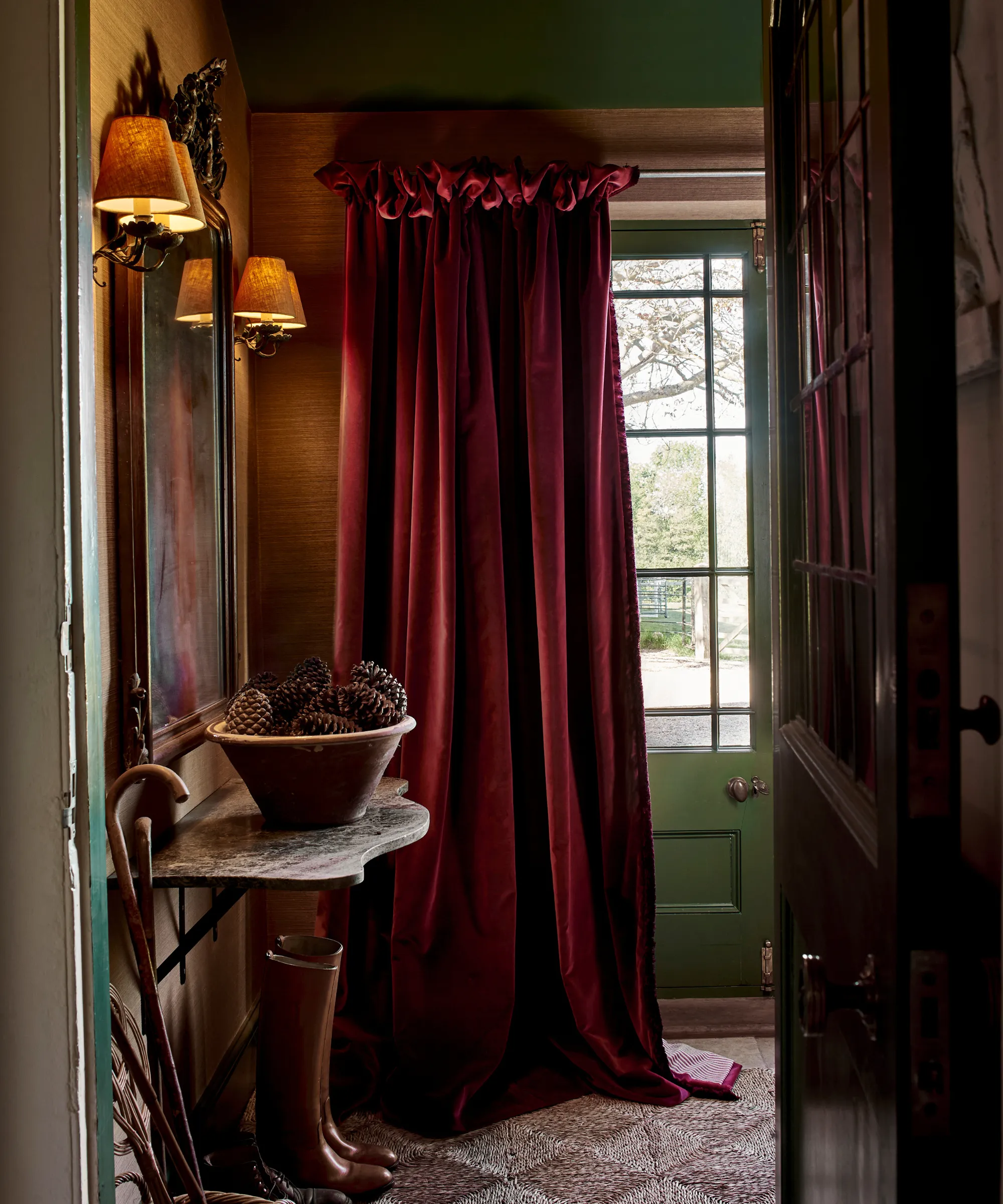 en entré med et dramatisk rødt fløjlsgardin et marmorbord med en skål fyrrekogler et kunstværk hængende op ad væggen med to lampetter på hver side og et par støvler på gulvet' loading='lazy' title=