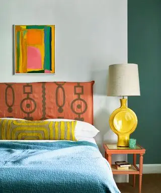 Quarto colorido com paredes pintadas de branco e verde, obras de arte abstratas coloridas acima da cama, cabeceira estampada e estofada laranja, cama com lençóis brancos, almofada estampada amarela, manta azul, abajur escultural amarelo com abajur creme na mesa de cabeceira laranja