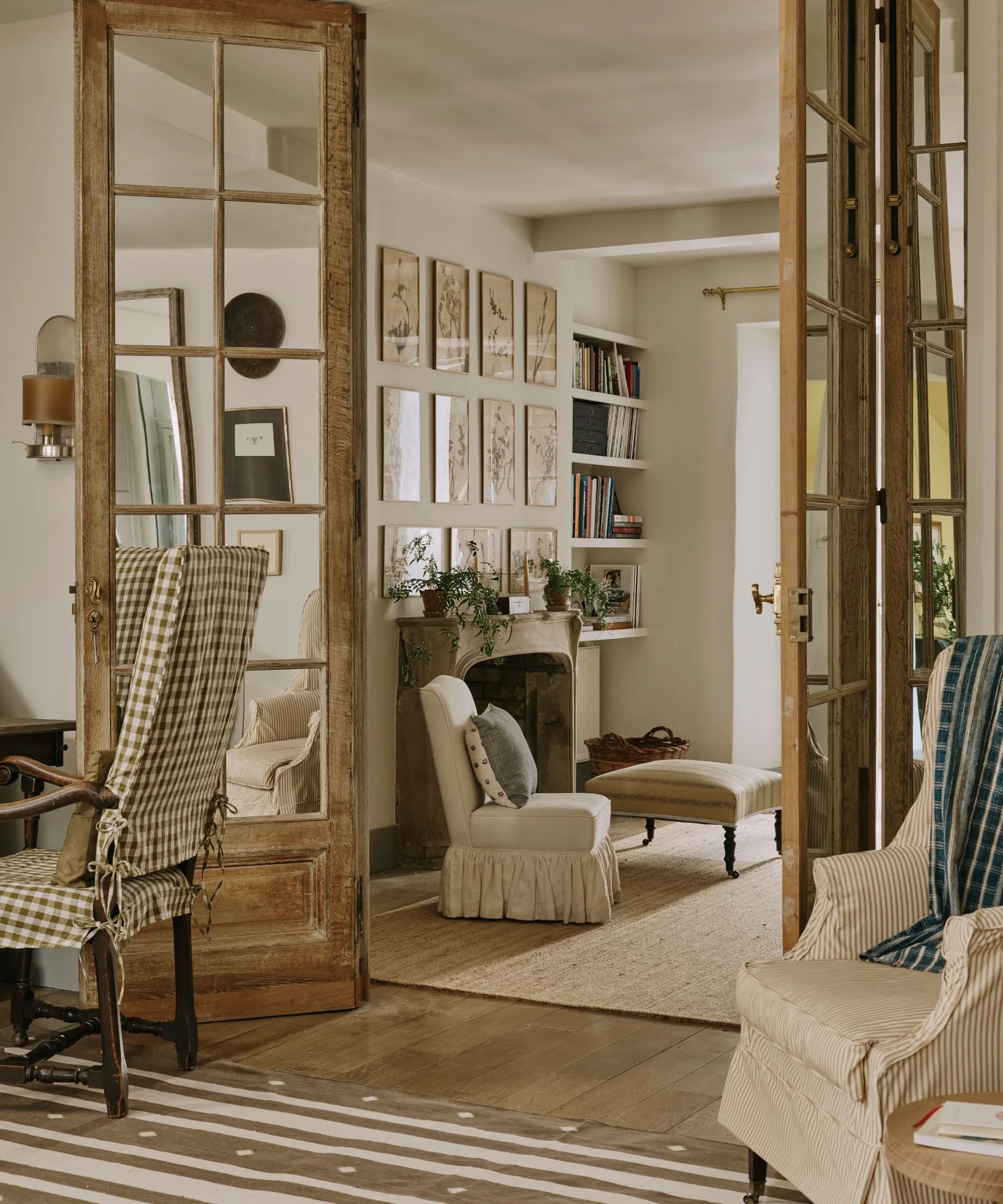 מבט דרך דלתות צרפתיות מעץ שעבר מזג אוויר אל חלל מגורים בגוון ניטרלי שליו. החדר כולל אח אבן, קיר גלריה של סקיצות בוטניות וריהוט נעים עם כיסוי החלקה כולל כורסה עם חצאית קפלים וכיסא ג'ינג'ם ירוק-לבן בחזית' loading='lazy' title=