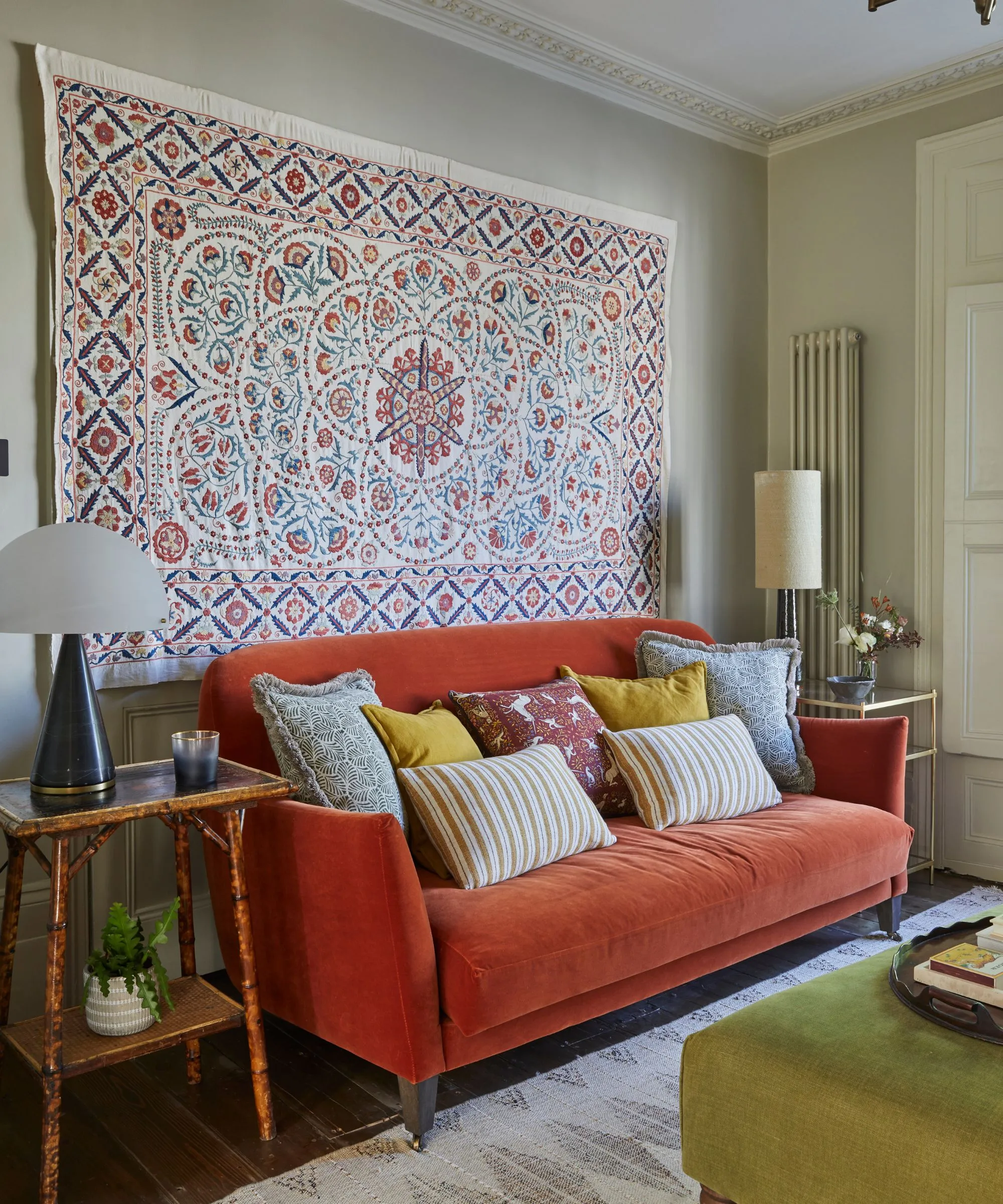 リビングルームには赤いソファがあり、その上に枕がたくさん置かれています。後ろの壁から大きなタペストリーがぶら下がっており、ソファの両側にはサイドテーブルがあります。' loading='lazy' title=