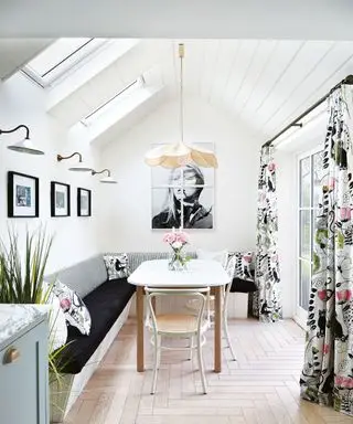 Lyst, hvitt kjøkken med panelt hvelvet tak, spisebord i tre med sittebenk, blomstergardiner, gulv i sildebeinstil, vegglamper, lavthengende pendel over bordet