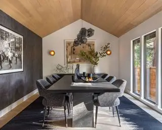 Spisestue med hvelvet tak, kledd i varmende tre, stort spisebord i svart marmor med polstrede spisestuestoler, moderne hengende lysarmatur, blått teppe på tregulv, grå strukturvegg med kunstverk