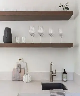 Nøytral kjøkkenplass med fokus på to trehyller plassert over kjøkkenvasken. Stilt på en minimalistisk måte med glass, dekorative vaser og plante. Benkeplate i naturlig kremstein med skjærebrett i marmor og tre, håndsåpe og sølvkran