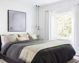 Quarto principal tranquilo com paredes e cortinas pintadas de branco, cama grande estofada em preto com roupa de cama e almofadas cinza, obras de arte acima da cama, pingentes suspensos de concreto