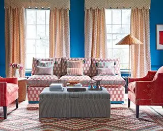 Sala azul com cortinas amarelas, sofá rosa e poltronas combinando, tapete estampado, pufe listrado.