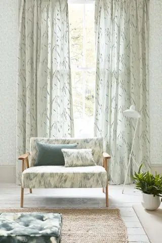 cortinas da sala com estampa de folhas e papel de parede claro, piso de madeira branca, tapete de coco, poltrona retrô estofada