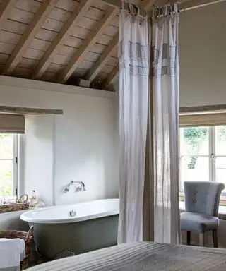 Quarto rústico rústico com vigas expostas no teto abobadado de madeira, cama com dossel de madeira e cortinas transparentes macias, banheira ao lado da janela