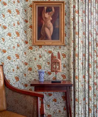 Close de papel de parede laranja e verde e cortinas combinando, foto emoldurada dourada, mesa lateral de madeira escura e poltrona