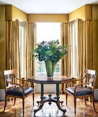 Sala de estar clássica com mesa e cadeiras de madeira escura, paredes pintadas de mostarda e cortinas combinando com sanefa na área da janela grande, vaso grande com flores na mesa