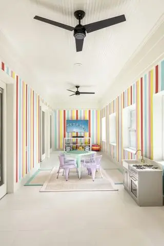 sala de juegos con paredes estilo rayas color caramelo, piso de baldosas blancas, silla y mesa de color pastel, dos alfombras, área de juegos en la cocina, juguetes y techo traslapado