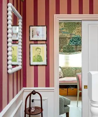 Papier peint à rayures rose clair et foncé, miroir de style bobine blanc, œuvres d'art, panneaux blancs, table d'appoint en bois, vue sur la pièce avec store à motifs verts et bleus