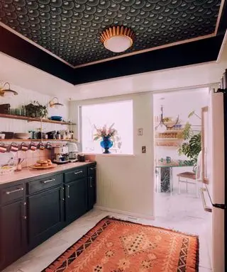 Cuisine blanche avec papier peint noir festonné au plafond, plafonnier doré, tapis à motifs roses au sol, armoires de cuisine noires, donnant sur l'espace salle à manger