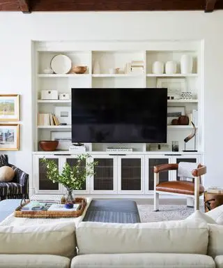 Sala de estar neutra com prateleiras altas decoradas, TV preta grande, sofá creme, obras de arte nas paredes, pufe, poltrona de couro