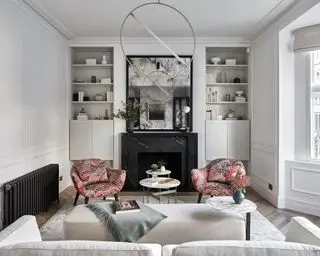 Stue med lysegrå vegger, karnapp, innebygd bod på hver side av pipe og peis. Grått teppe og sofa og oransje mønstrede lenestoler.