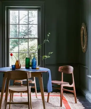 Un'idea cromatica per la sala da pranzo con pareti verde smeraldo, specchio con cornice in rattan, sedie e tavolo in legno dal design moderno e tappeto in tessuto naturale