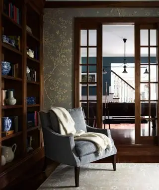 Estudio de oficina en casa oscuro con pisos de madera oscura, puertas francesas y paredes revestidas con estanterías de caoba y un cómodo sillón.