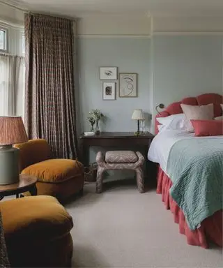 Dormitorio pintado de azul claro con una cama rosa, sillones de terciopelo mostaza y muebles de madera oscura.