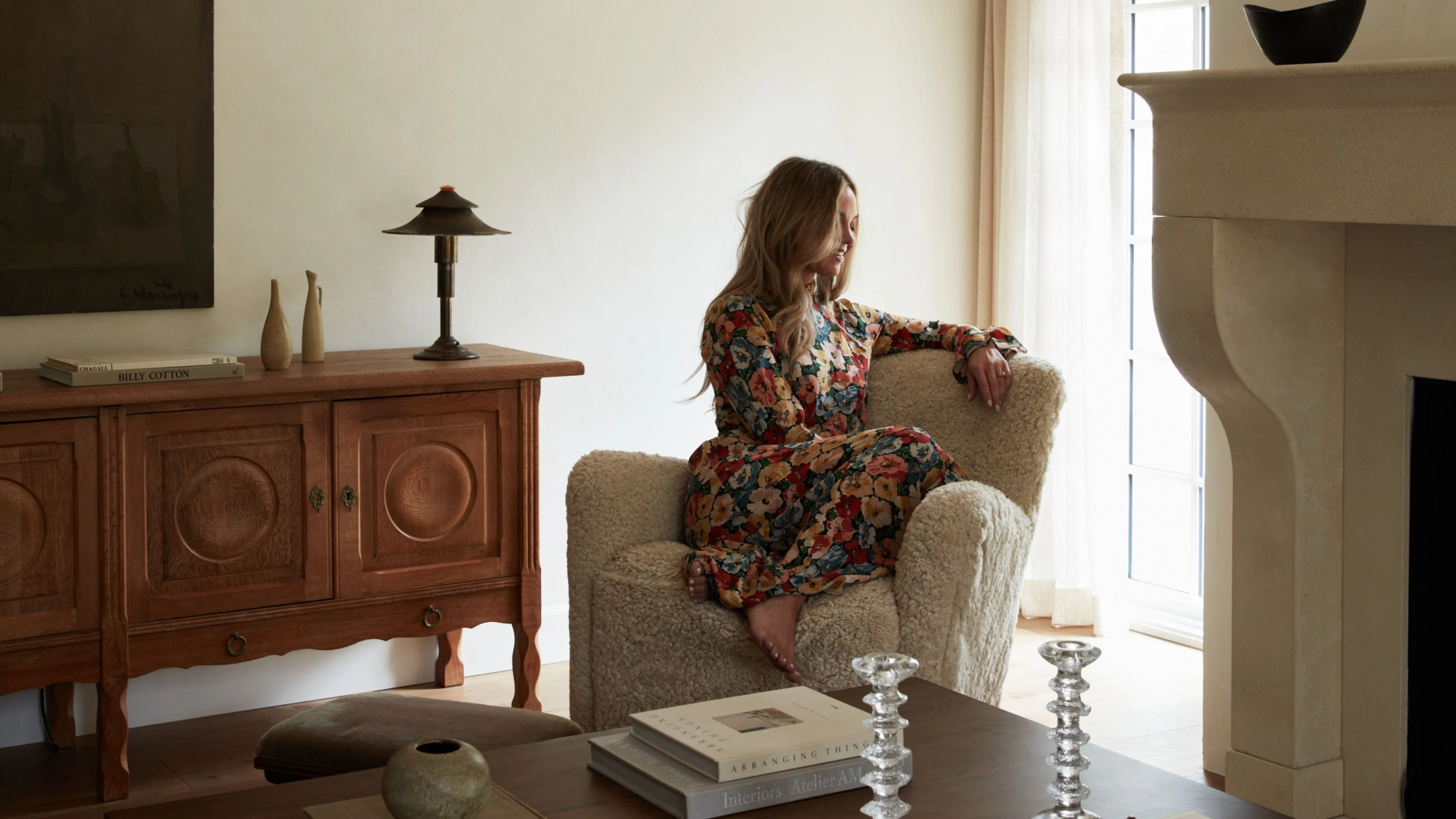 インテリアデザイナーのモリー・キッドさんは、木製の真鍮やセラミックのオブジェを集めた温かみのあるミニマルなオレゴン州のリビングルーム内で、メイデン・ホームのムートンのアクセントチェアの上にポーズをとっている。' loading='lazy' title=
