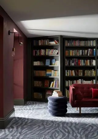 sala de estar no subsolo com carpete cinza estilo zebra, banquinho redondo cinza, sofá de veludo vermelho clarete, estantes, paredes vermelho clarete, teto branco, apliques