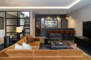 sala de estar / sala de jogos no porão com grande seção de veludo castanho-avermelhado, TV, bar com bancos de bar, mesa de console preta, banquinho de couro, portas abertas para o andar de cima