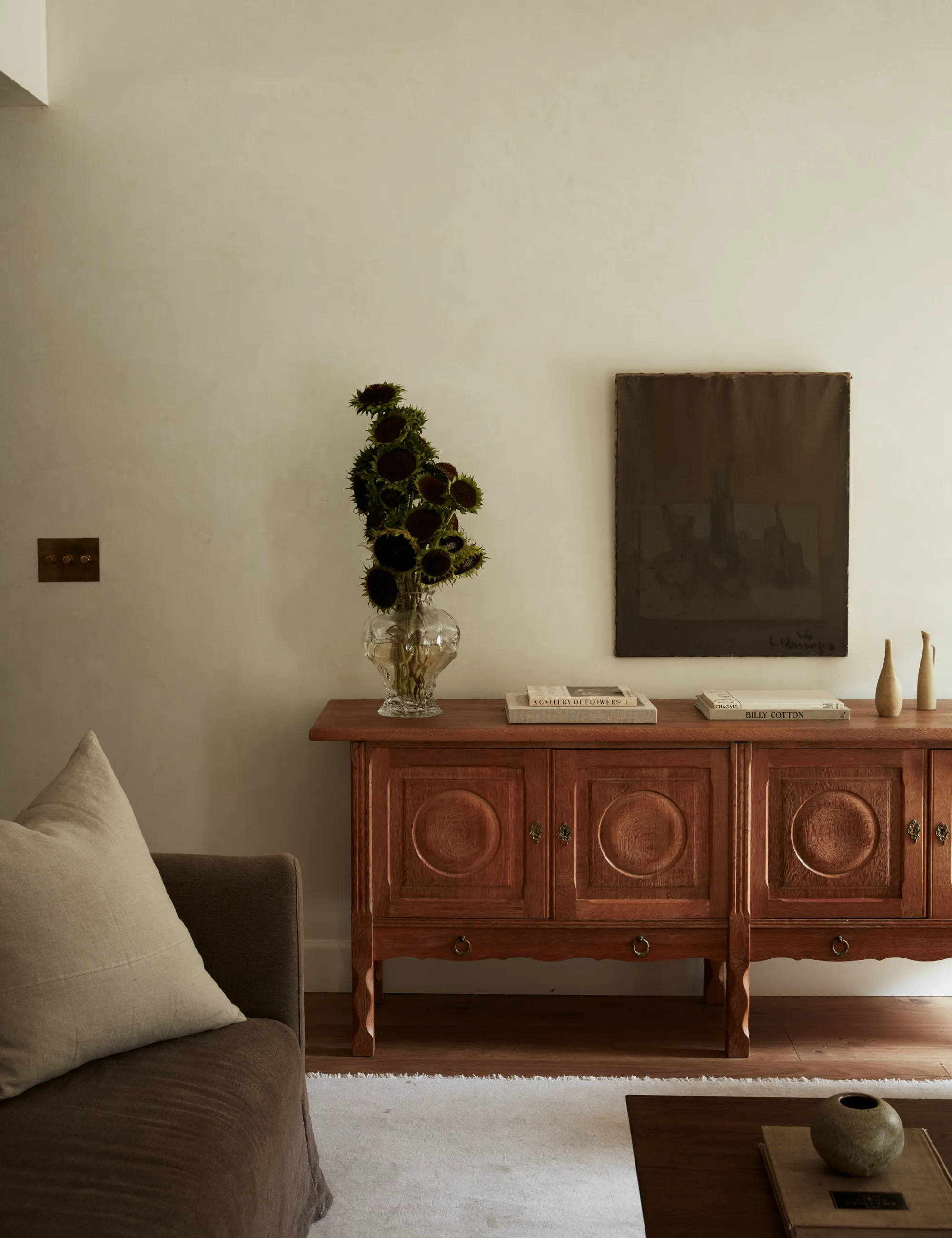 Aparador de madera de mediados de siglo con un jarrón transparente con girasoles, un tapiz de pared apagado y varios objetos coleccionados.' loading='lazy' title=