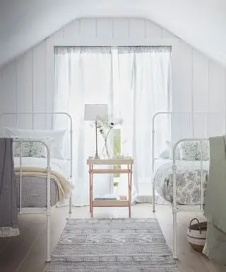 Ein helles und luftiges weißes Schlafzimmer mit zwei Einzelbetten aus Metall mit Toile-Vorhängen und weißen Wänden