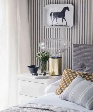 Ein Beispiel für Schlafzimmerbeleuchtungsideen mit einer übergroßen Tischlampe aus Glas und Gold vor einer gestreiften Tapete