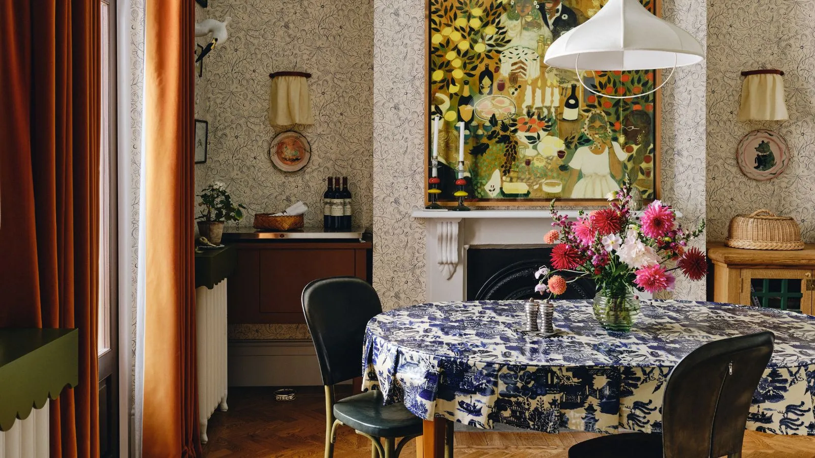 Sala de comedor de estilo máximo escandi con paredes con papel tapiz una gran chimenea con arte popular colgado sobre cortinas de terciopelo de naranja oscura Una mesa de comedor con mantel estampado azul y blanco y una luz colgante de tela blanca luz' loading='lazy' title=