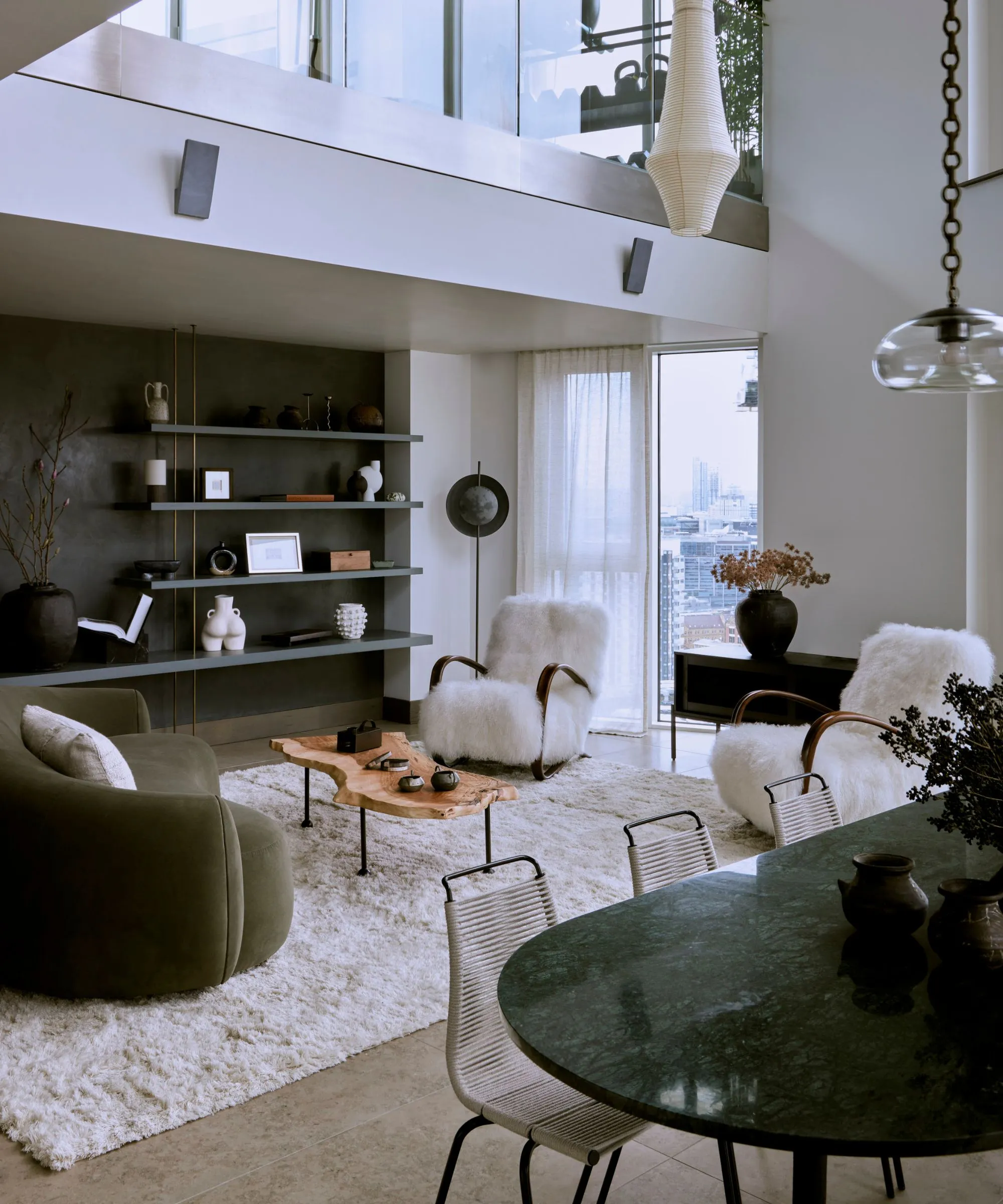 Acogedora área moderna de sala de estar de planta abierta con una mesa de comedor configurada con una mesa de mármol verde sillas modernas una gran sala de estar con una alfombra blanca esponjosa con un sofá curvo de terciopelo verde y sillas de brazo de piel de oveja' loading='lazy' title=