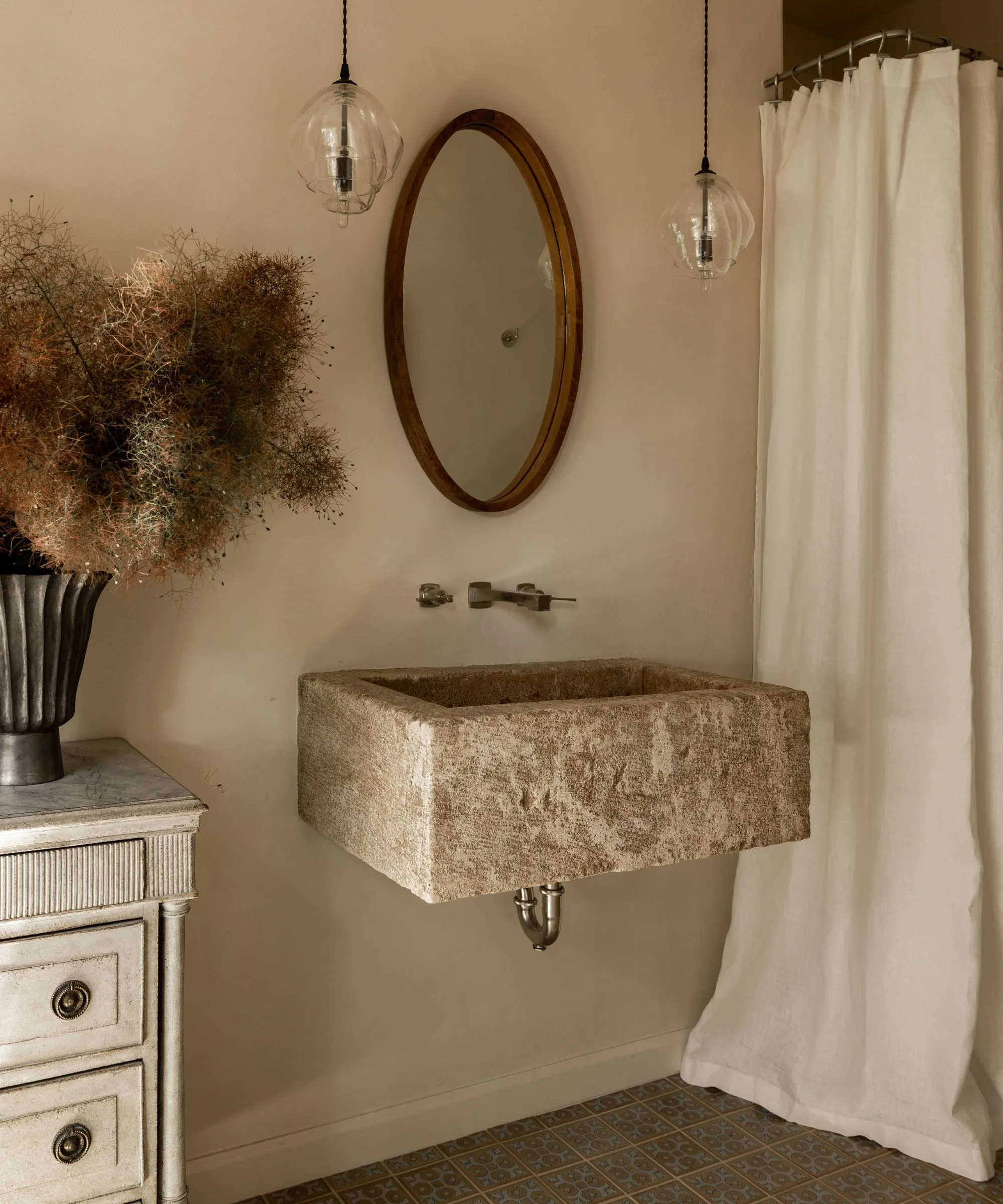 Baño pequeño neutro tibio con una cortina de lino blanca de piedra fregadero flotante espejo de madera y un tocador antiguo con un jarrón con ramas' loading='lazy' title=