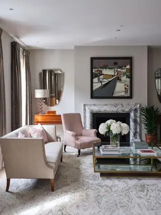 Wohnzimmer mit hellrosa Sofa und Sessel neben Kamin mit Marmorverkleidung, hellgrauem Teppich und verspiegeltem Couchtisch