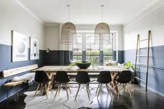 sala da pranzo con pareti grigio ardesia dipinte a metà altezza, pareti superiori e soffitto dipinti in pietra, pavimento in legno scuro, tavolo da pranzo rustico, sedie Eames nere, opere d'arte, tappeto con animali, paralume in rattan, panca