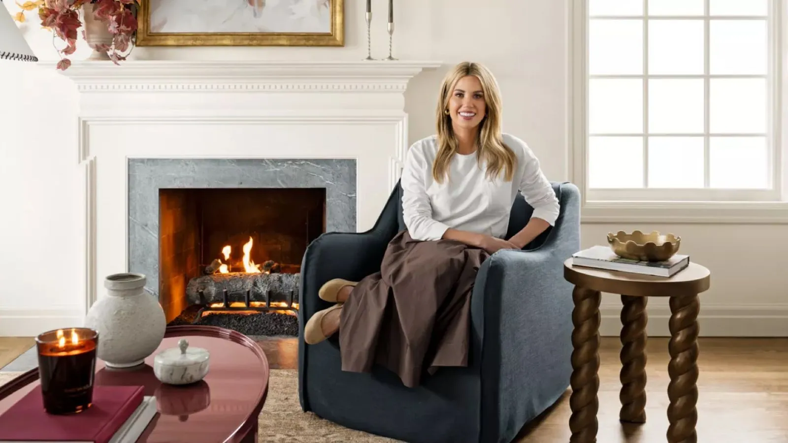 Designer Shea McGee fotograferet til sin Target 2025-kollektion sad på en marineblå lænestol med slipbetræk ved siden af en pejs med et drejet sidebord i træ og et lakeret rødt sofabord' loading='lazy' title=