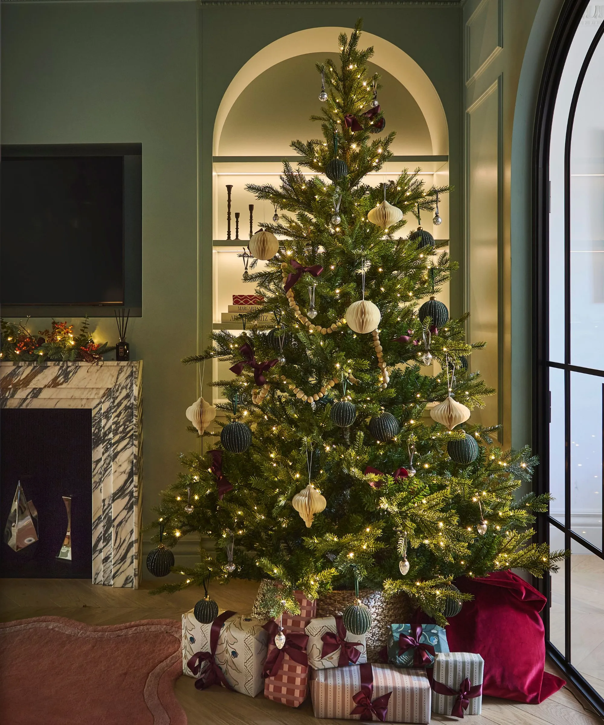 סלון ויקטוריאני ירוק מושתק עם אח משיש כוננית ספרים מקושתת טלוויזיה, הכל מקושט לחג המולד עם עץ גדול מואר ומתנות עטופות' loading='lazy' title=